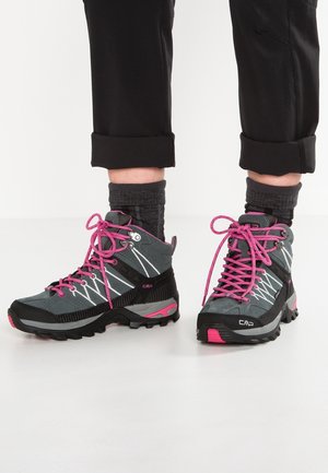 Persoon draagt zwarte broek met omslag, donkergrijze wandelschoenen met roze veters en accenten, en donkergrijze sokken op een witte vloer.