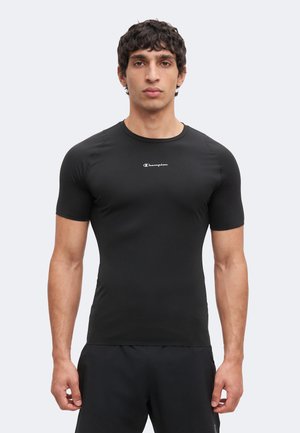 Jeune homme face à l'objectif, portant un t-shirt noir Champion ajusté et un short noir sur un fond clair uni.