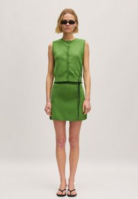 Groene gebreide mouwloze jurk met knoopsluiting en een zwarte trekkoord taille. Soepele textuur, lengte boven de knie, vergezeld van zwarte sandalen.