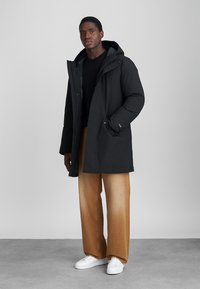 Μαύρη επενδυμένη parka με κουκούλα, διαθέτοντας μπροστινό φερμουάρ και κλείσιμο με κουμπιά, πλαϊνές τσέπες και χαλαρή εφαρμογή, συνδυασμένη με ανοιχτό καφέ παντελόνι σε φαρδιά γραμμή.