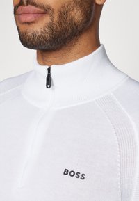 Bela zip-up trenerka z visokim ovratnikom, ki ima rebrasto teksturo in črn logo "BOSS" na prsih.