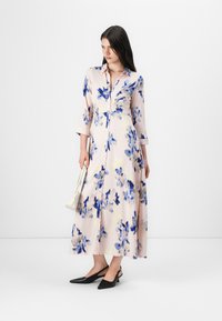 En kvinna i en blommig rosa klänning med blå detaljer står med en hand som håller en liten vit handväska, iförd svarta slingbacks.