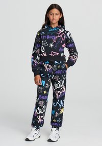 Felpa nera con cappuccio e pantaloni abbinati con motivi colorati in stile graffiti. Il materiale è morbido; il design presenta un top corto e una cintura elastica.