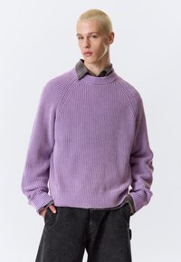 Pull violet côtelé avec un col montant, des manches raglan et une texture en relief, superposé sur une chemise à carreaux et associé à un pantalon foncé.