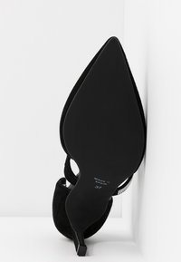 Chaussure à talon haut noire pointue vue de la semelle avec "Made in Spain" et taille 37 visibles, posée contre un mur blanc en angle.