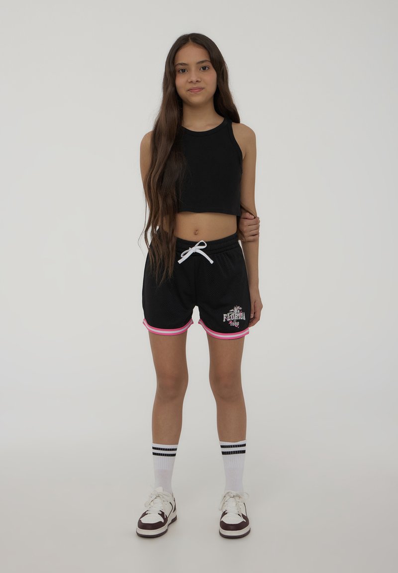 Canotta nera corta e pantaloni corti in rete neri con rifiniture rosa. Stampa caratteristica sui pantaloni. Calzini bianchi con strisce nere e sneakers.