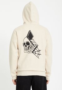 Beige hoodie met een grote zwarte afbeelding van een schedel, trappen en een spinnenweb op de achterkant. Inclusief ribgebreide manchetten en tailleband.