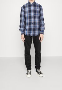 Camicia a quadri blu scuro e blu, a maniche lunghe, abbinata a pantaloni neri e sneakers nere con suole bianche. Polsini regolabili.