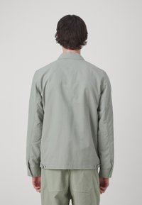 Cămașă lungă cu mânecă, de culoare verde deschis, cu guler, textură netedă și nasturi laterali, purtată peste pantaloni asortați, prezentând un design minimalist.
