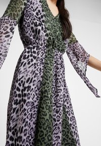 Průhledné šaty s tygřím vzorem s fialovými a zelenými leopardími motivy, s výstřihem do V a dlouhými, splývavými rukávy. Detail s elastickým pasem.