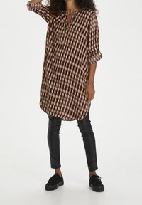 Printed tunic with a brown, pink, and black geometric pattern; lightweight fabric, collarless, three-quarter sleeves, relaxed fit. Black leather pants and shoes.