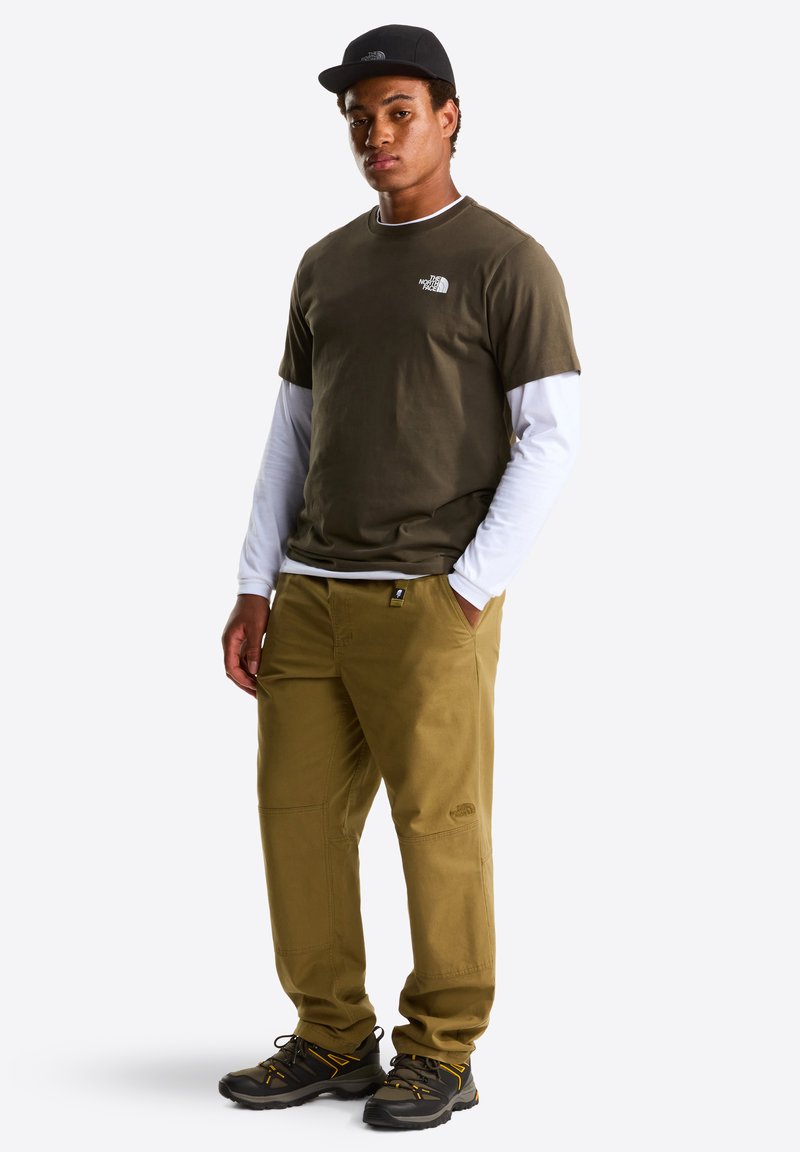 Olive green t-shirt with logo, layered under a white long-sleeve, paired with tan pants. Black cap and rugged black shoes with yellow accents.