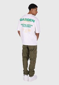 Hvid t-shirt med grønne og gule grafik på ryggen; parret med olivengrønne cargo-bukser og hvide sneakers.
