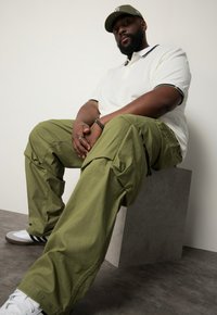 Beige polo shirt met contrasterende kraag, olijfgroene cargobroek met zijzakken, en witte sneakers met zwarte strepen, zittend op een grijs pedestal.