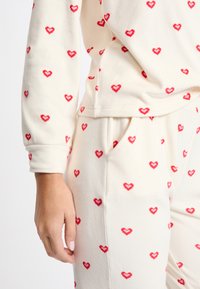 Valkoiset fleece-setti punaisilla sydänkuvioilla. Yläosa on rento malli ja ribatut hihansuut; alaosassa on taskut ja joustava vyötärö.