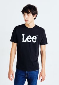 Melna kokvilnas T-kreklu ar izteiktu balto "Lee" logotipu. Ar apaļu kakla dizainu un īsām piedurknēm. Valkāts ar ziliem džinsu biksēm.