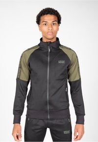 Chaqueta negra con cierre frontal y paneles en verde oliva en los hombros, logo "WRONG FRIENDS" en el pecho y las mangas, fabricada con un tejido texturizado y flexible.