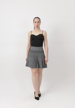 Kvinde stående mod en hvid baggrund iført en sort tætsiddende ærmeløs top, sort-hvid hundetandsnederdel og sorte højhælede sko.