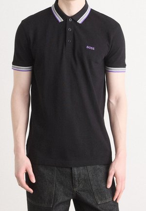 Mężczyzna w czarnej koszulce polo z fioletowo-białym obszyciem kołnierza i rękawów, logo „BOSS” na lewej piersi, w ciemnych dżinsach.