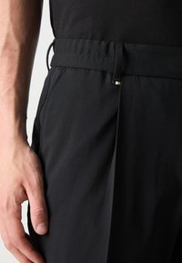 Pantaloni negri cu un față plată, buzunare laterale și o talie elastică cu un mic accent metalic. Materialul pare neted și ușor.