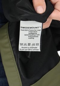 Swedemount-jackans etikett med en svart tygetikett med vit text, som anger 100 % polyester skal och skötselråd.
