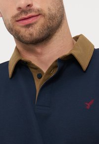 Άνδρας με ελαφριά γενειάδα που φοράει μπλε σκουρό πουκάμισο πόλο με καφέ γιακά από κορντόρι και μικρό κόκκινο κεντητό λογότυπο πουλιού στο στήθος.