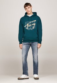 Sudadera de color azul verdoso con un logo gráfico en el frente, combinada con jeans azules desgastados y zapatillas blancas. La sudadera cuenta con cordones y un bolsillo tipo canguro.
