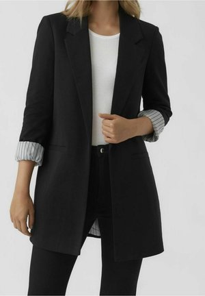 Kobieta ubrana w czarną marynarkę z podwiniętymi, pasiastymi mankietami, na białej, prążkowanej koszulce i czarnych spodniach, stojąca na jednolitym tle.