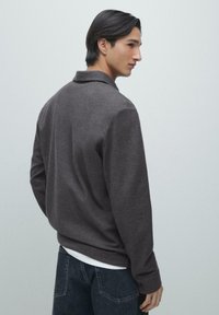 Donker antracietgrijs shirt met lange mouwen en kraag, gemaakt van getextureerde stof; gedragen over een wit ondershirt en gecombineerd met donkere jeans.