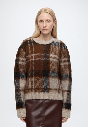 Femme aux cheveux blonds raides portant un pull à carreaux marron, beige et noir et une jupe en cuir marron foncé sur un fond uni.