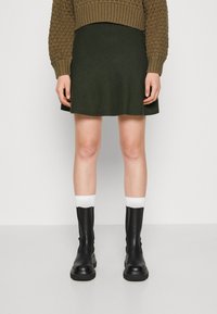 Jersey de color verde oliva con un patrón texturizado, combinado con una minifalda verde oscura. Botines de cuero negros y calcetines de punto blancos.