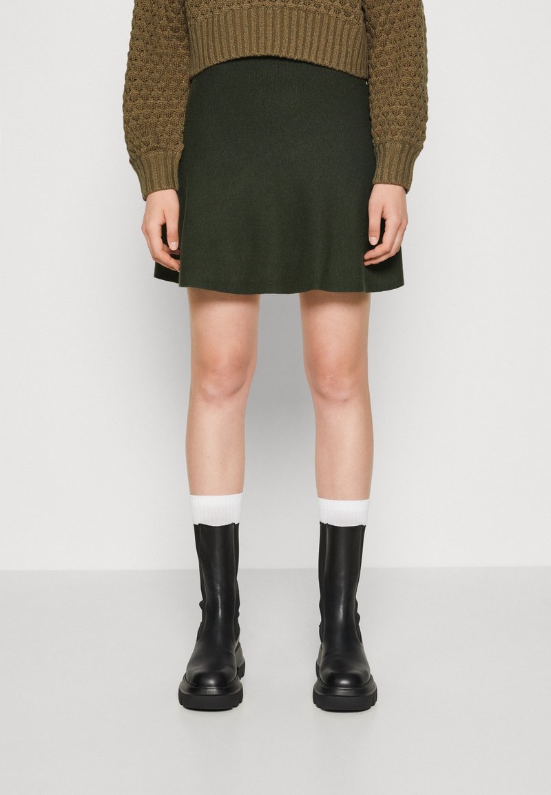 Jersey de color verde oliva con un patrón texturizado, combinado con una minifalda verde oscura. Botines de cuero negros y calcetines de punto blancos.