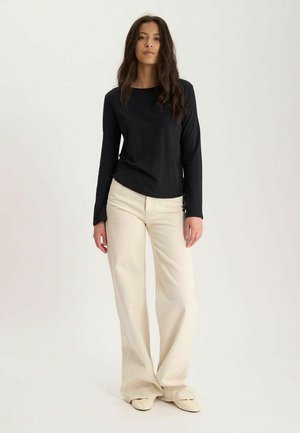 Femme aux longs cheveux foncés portant un haut noir à manches longues, un pantalon beige à jambes larges et des chaussures beiges sur un fond uni.