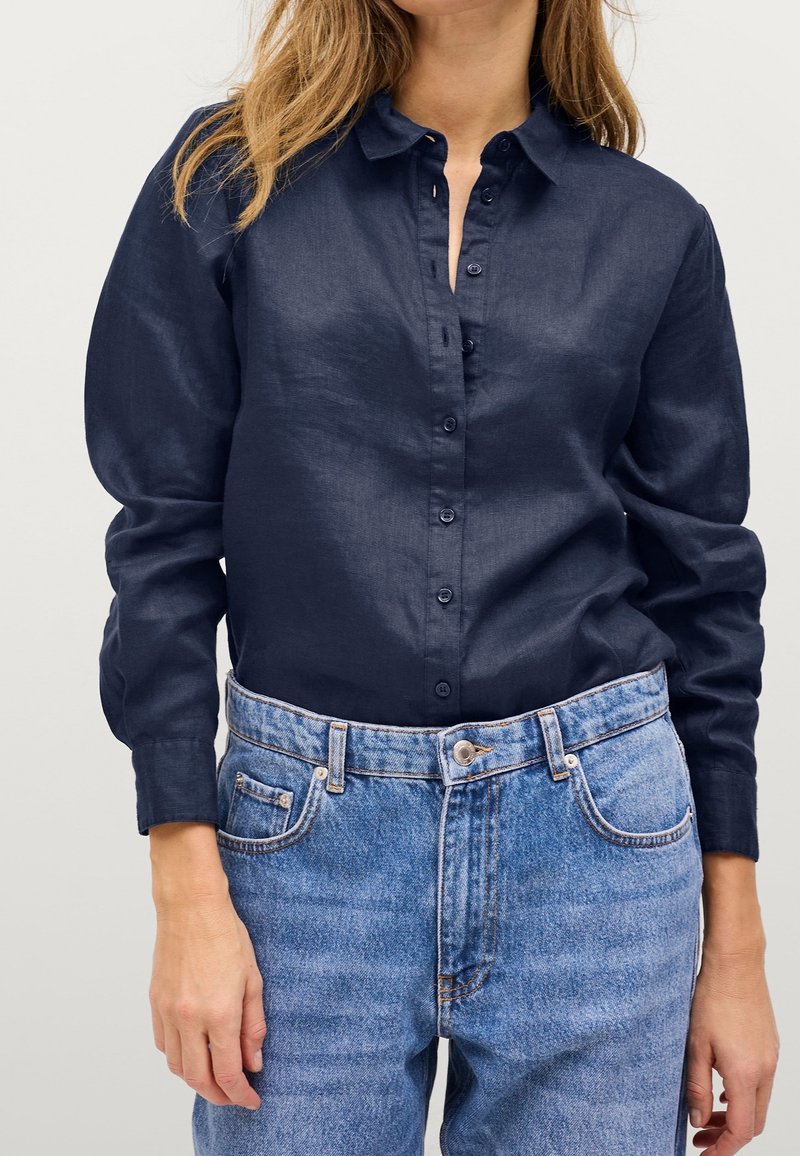 Marinblå, långärmad skjorta med knappar i slät tyg, med en klassisk krage och knappar fram, parat med ljusblå jeans.