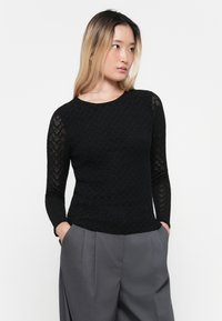 Black textured long-sleeve top featuring a round neckline and patterned sheer sleeves, paired with gray high-waisted trousers.