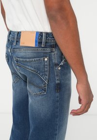 Σκούρο μπλε τζιν με απαλό υφή, πέντε τσέπες και μια καφέ δερμάτινη ετικέτα με μπλε λεπτομέρειες στη ζώνη.