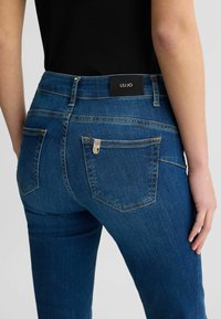 Jeans in denim blu scuro con vita alta, caratterizzati da cuciture gialle, etichetta con logo sul retro e due tasche posteriori con rivetti decorativi.