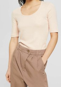 Světle růžové žebrované tričko s kulatým výstřihem a krátkými rukávy, spárované s béžovými kalhotami s vysokým pasem, které mají přední záhyby a boční kapsy.