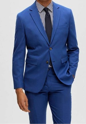 Mann trägt einen maßgeschneiderten, leuchtend blauen Anzug mit einem gestreiften Hemd und einer einfarbigen schwarzen Krawatte, eine Hand in der Tasche, steht vor einem schlichten Hintergrund.
