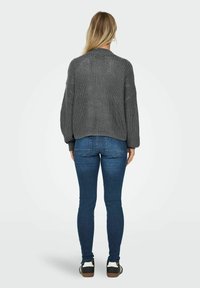 Siva pletena jopica z teksturiranim zaključkom, v kombinaciji s prilegajočimi modrimi kavbojkami in črnimi supergami z belimi detajli. Pogled od zadaj.