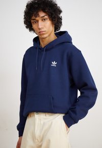 Marineblauwe hoodie van een katoenmix, met een kangoeroezak, een trekkoordcapuchon en een wit Adidas-logo op de borst. Het model draagt een crème broek.