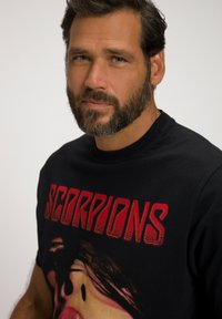 Černé tričko s červenou grafikou a nápisem "SCORPIONS" společně s ilustrací výrazu obličeje. Tkanina vypadá hladce a přiléhavě.