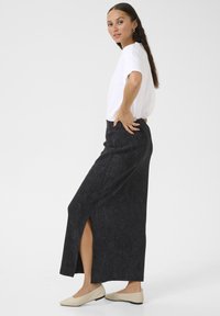 Black, high-waisted, long skirt with a side slit, made of textured fabric. Paired with a white, short-sleeve t-shirt and light shoes.
