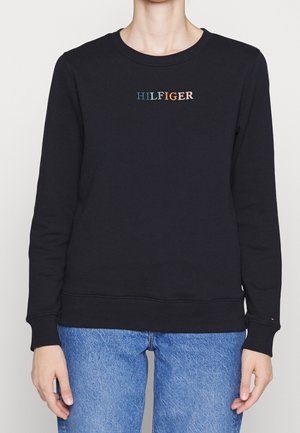 Osoba w czarnej bluzie z okrągłym dekoltem z wielokolorowym napisem „HILFIGER” i niebieskich dżinsach, stojąca na tle jasnego, jednolitego tła.