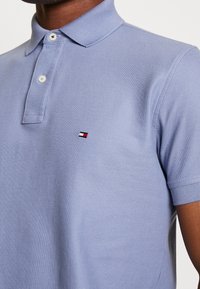 Ljusblå polo skjorta tillverkad av texturerad tyg. Har en klassisk krage, två knappslå och en liten logotyp i röd, vit och svart.