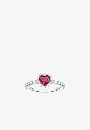 Bague en argent ornée d'une pierre précieuse rouge en forme de cœur, entourée d'un motif rayonnant avec des accents blancs et un anneau texturé.