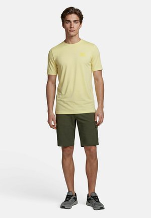Jonge man staat, draagt een lichtgeel T-shirt met korte mouwen, donker groene cargoshorts en grijze en zwarte sneakers.