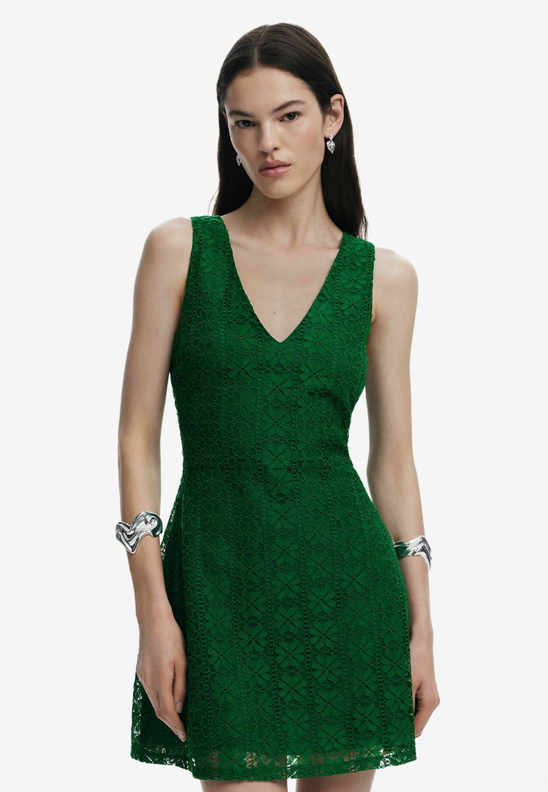 Grünes Spitzenkleid mit V-Ausschnitt, figurbetontem Oberteil und ausgestellt Rock. Detailliertes florales Muster und strukturierter Stoff. Silbernes Armreif am Handgelenk.