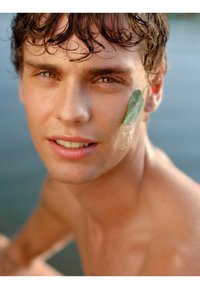 Close-up portret van een jonge man met nat, golvend bruin haar. Een groene blad ligt tegen zijn wang, waardoor zijn lichte huid en bruine ogen benadrukt worden.