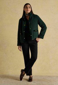 Veste en polaire verte avec des boutons-pression dorés, deux poches avant, pantalon noir et chaussures brunes avec un accent en chaîne dorée.